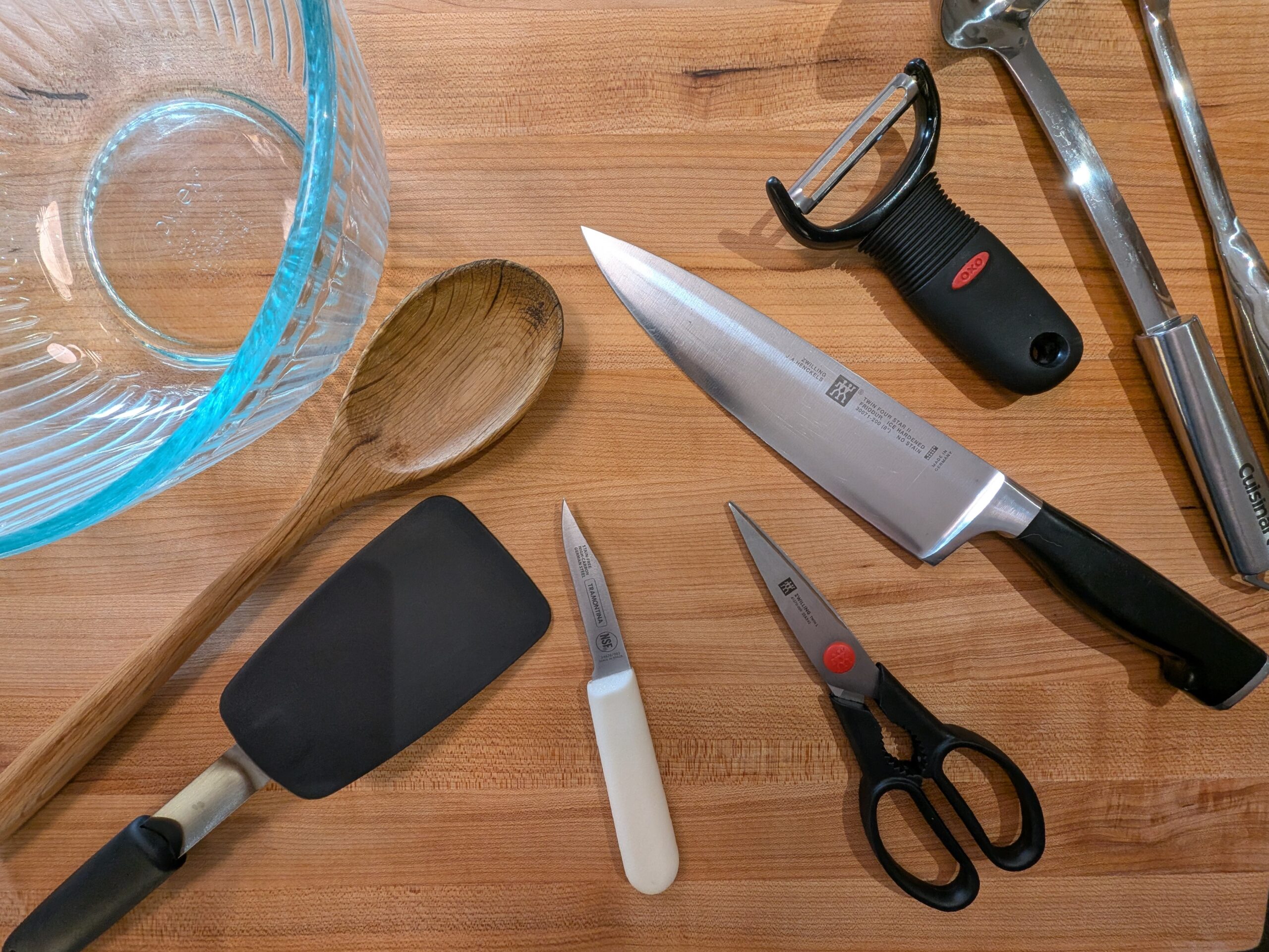 An array of kitchen equipment including a chef's knife, a pair of scissors, a y-peeler, a paring knife, a glass mixing bowl, a wooden spoon, and a silicone spatula